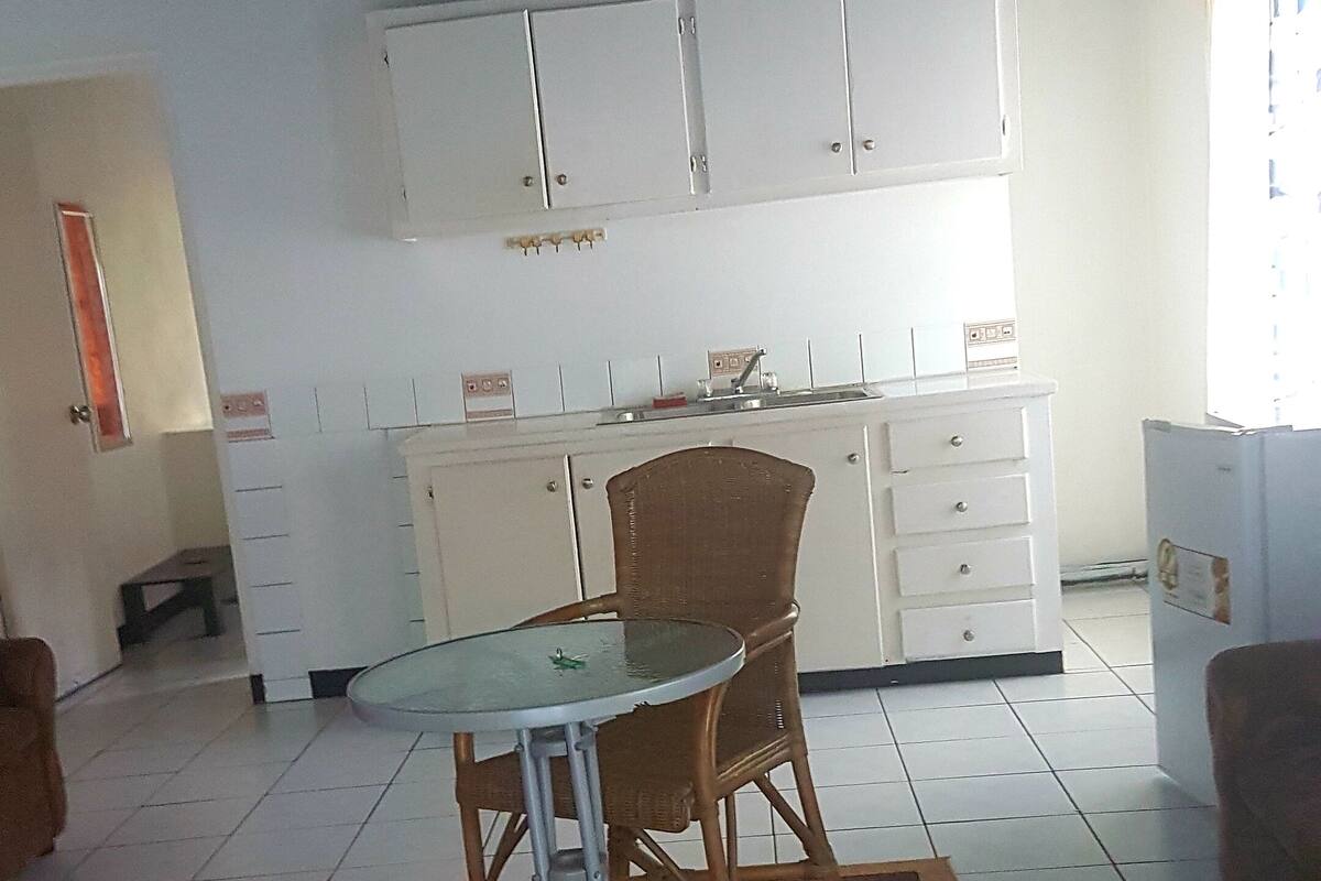 A dining area with a glass table and wicker chairs, a kitchen with white cabinets, and a tiled floor.