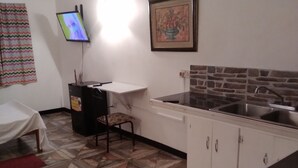 A compact kitchen with a tiled floor, a dining area with a table and chairs, a TV mounted on the wall, and a framed picture above the sink. - Honiiwell rooms and suites (St. George's)