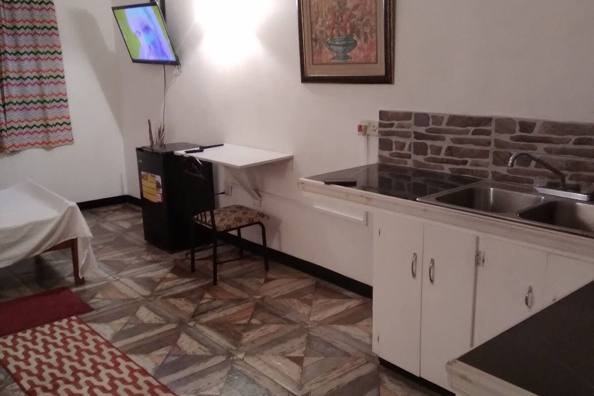 A compact kitchen with a tiled floor, a dining area with a table and chairs, a TV mounted on the wall, and a framed picture above the sink.