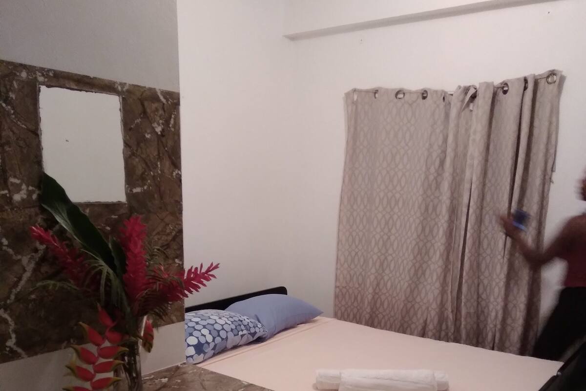A bedroom with a bed, a nightstand, a mirror, a vase with flowers, a patterned rug, and a window with curtains.