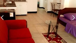 A living room with a red sofa, a glass coffee table, and a purple sofa. There is a kitchen in the background with white cabinets and a tiled backsplash. - Honiiwell rooms and suites (St. George's)