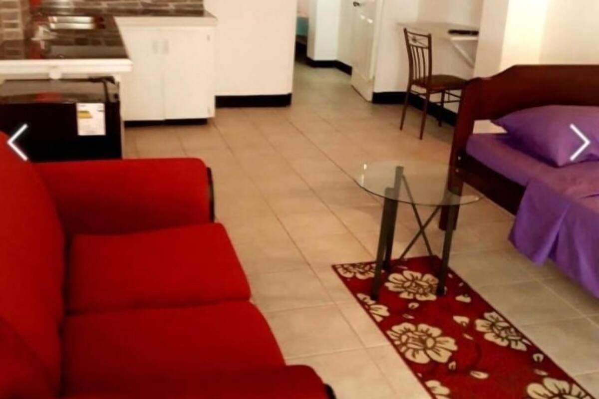 A living room with a red sofa, a glass coffee table, and a purple sofa. There is a kitchen in the background with white cabinets and a tiled backsplash.