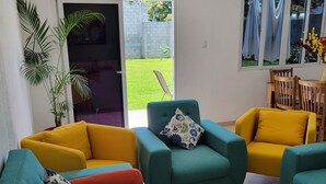 Un salon avec des canapés aux couleurs turquoise et jaune, une grande plante en pot et une vue sur l’extérieur.