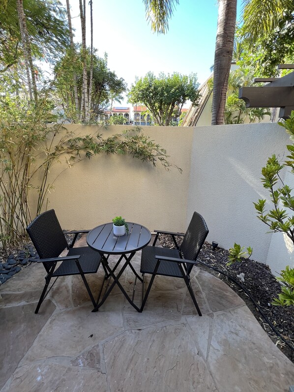 A patio with a round table and four chairs, surrounded by plants and a beige wall. - Brand New Luxury Maui Kamaole Condo Grd Flr D112 (Kihei)