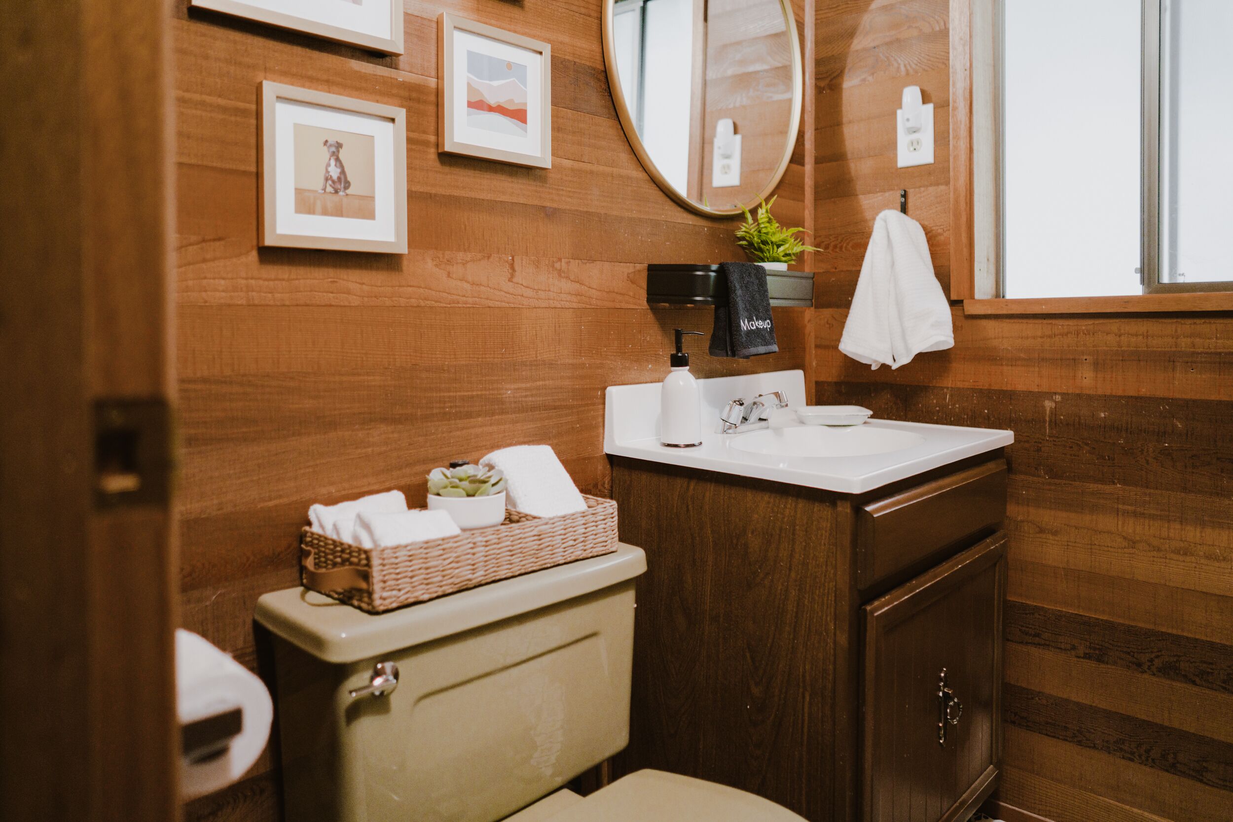 A wooden-paneled bathroom with a toilet, sink, mirror, and framed pictures on the wall.