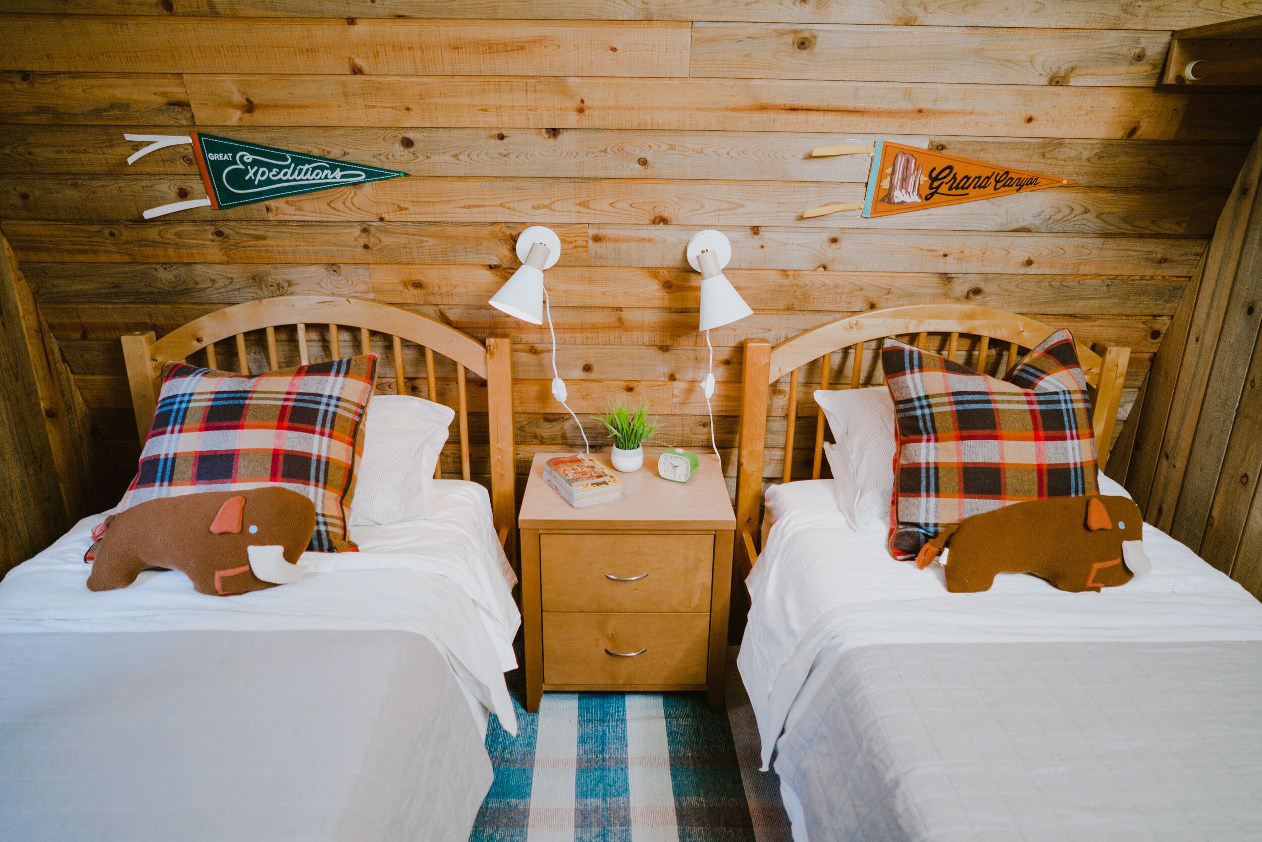 A cozy cabin-style room with two wooden beds, plaid pillows, a nightstand with a lamp, and a small plant.