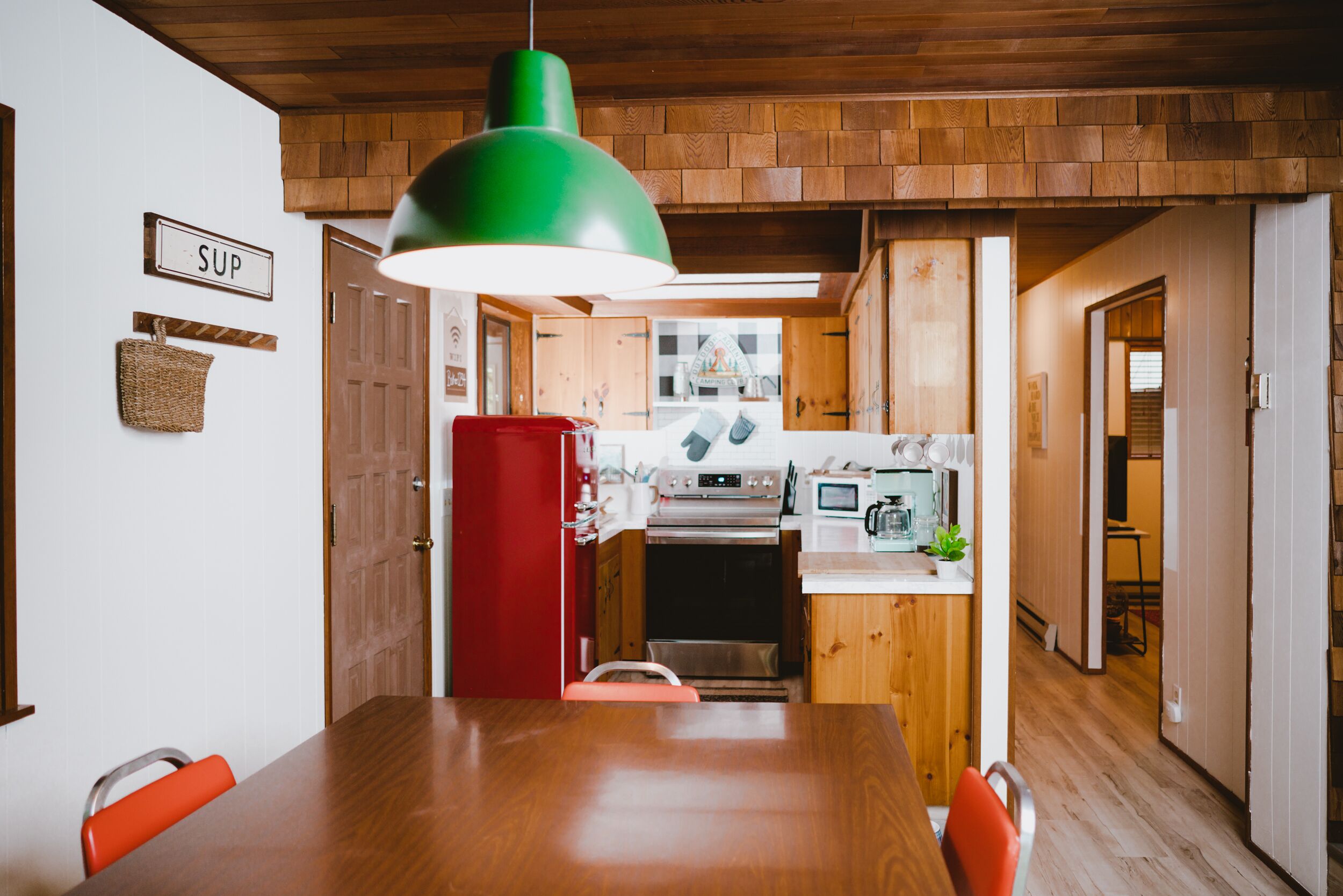 A wooden dining table with chairs, a red refrigerator, and a kitchen area with a microwave and oven.