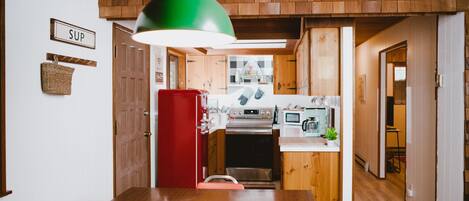 A wooden dining table with chairs, a red refrigerator, and a kitchen area with a microwave and oven.
