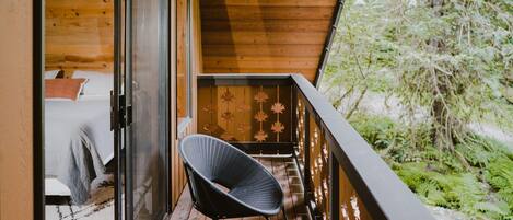 A wooden balcony with a chair, a bed with a blanket, and a door leading to a room.
