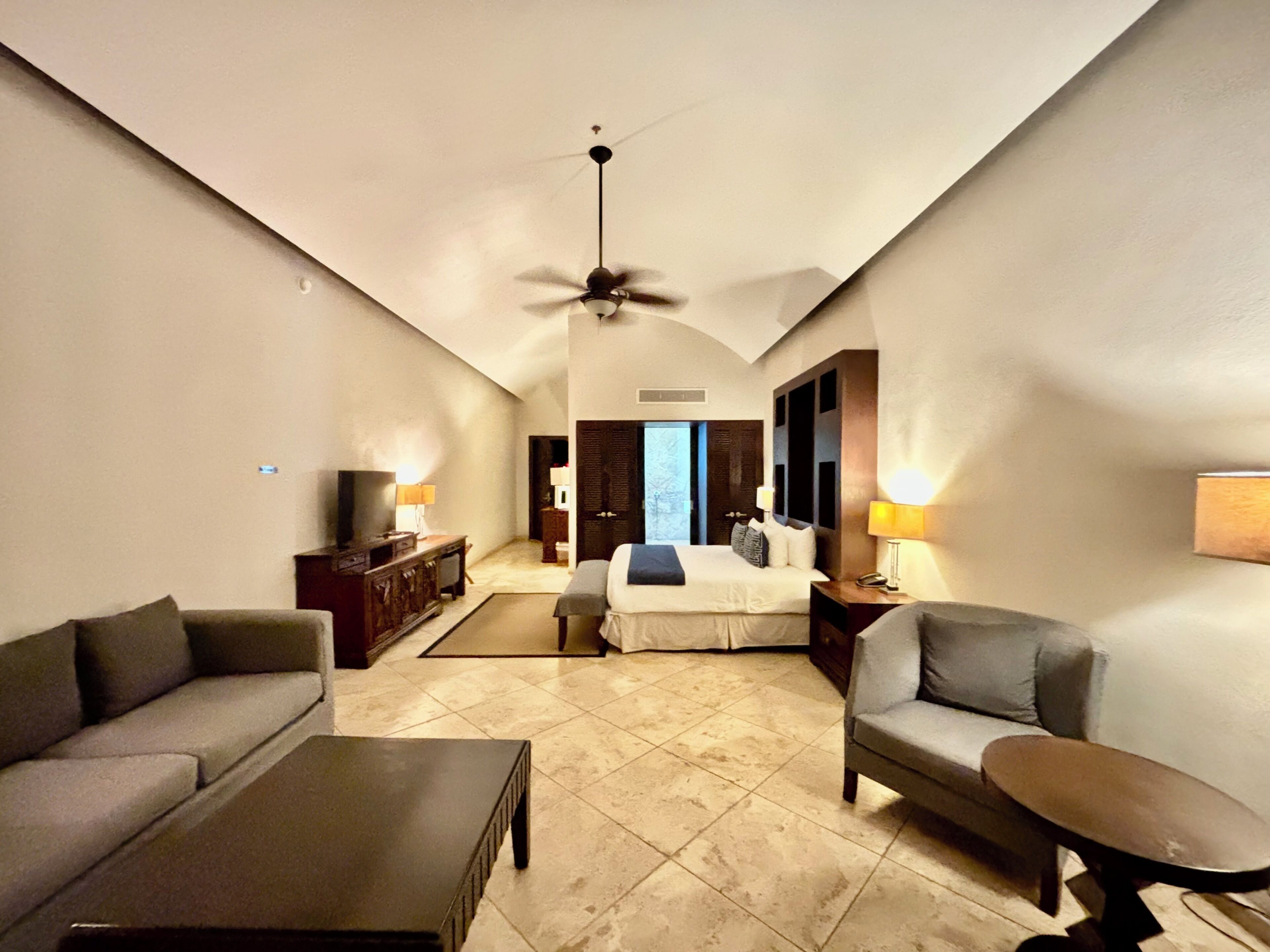 A hotel room with a bed, two armchairs, a coffee table, a TV, and a ceiling fan.