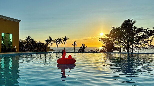 Una piscina con un flotador rojo inflable, palmeras y vistas al atardecer.