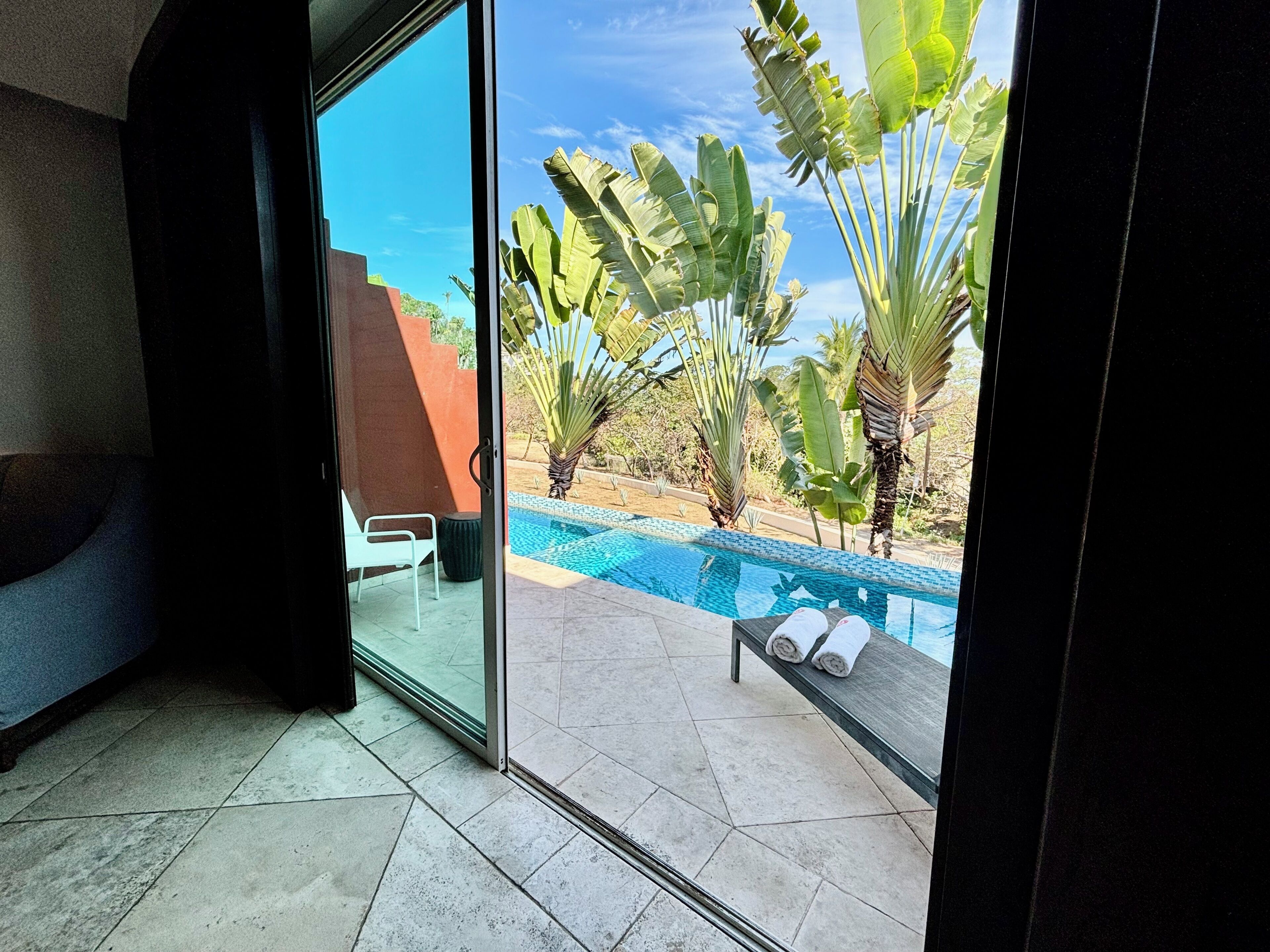 A room with a glass door leading to a pool area, featuring a bench with towels, a white chair, and a view of a pool and tropical plants.