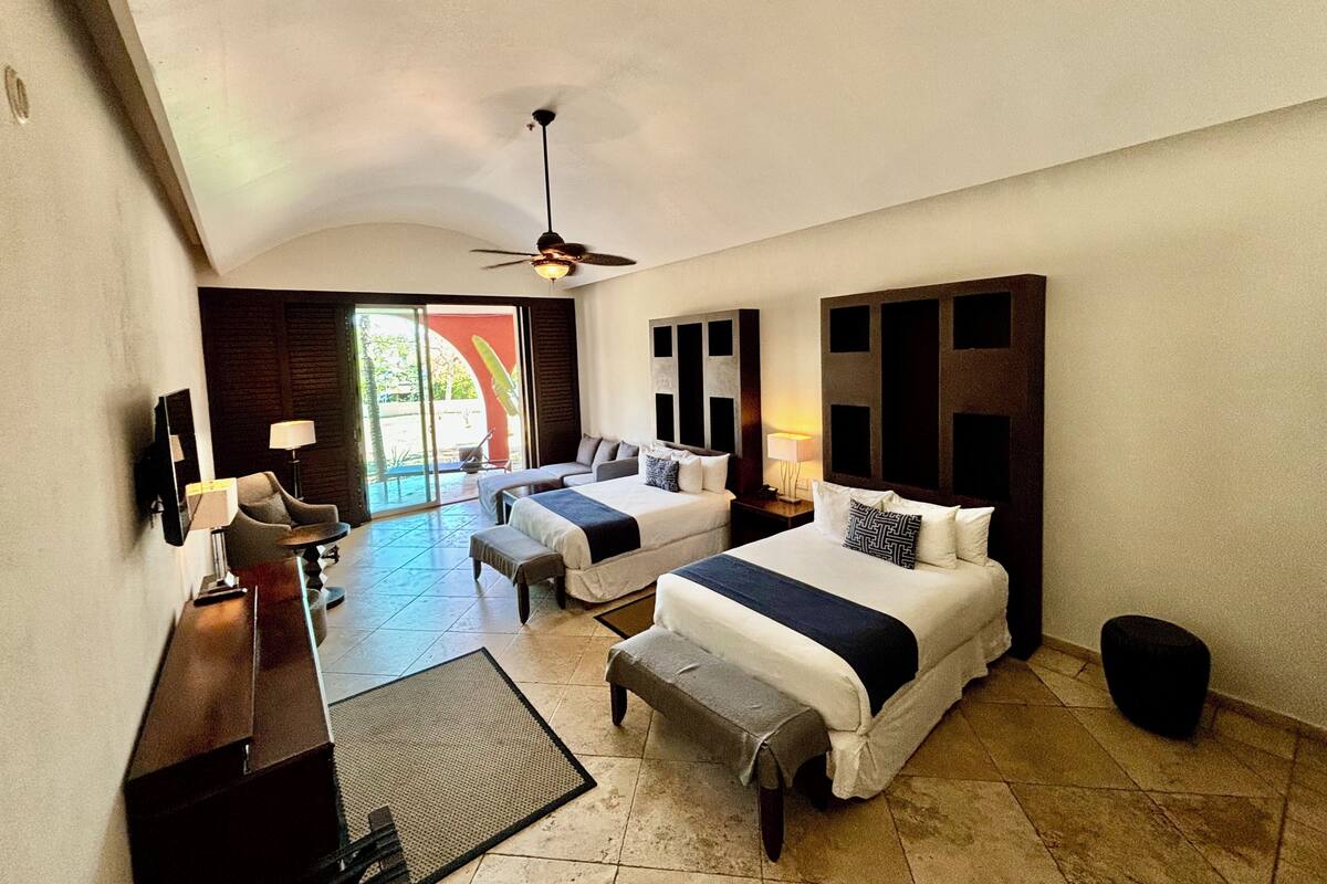 Une chambre d’hôtel avec deux lits, un bureau et une chaise. Elle est équipée d’un ventilateur de plafond, d’une télévision et d’une fenêtre donnant sur l’extérieur.