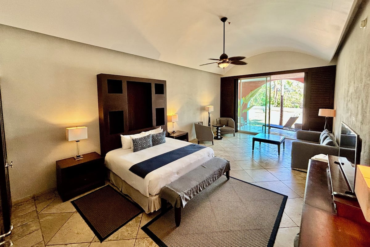 Une chambre d’hôtel avec un grand lit, un ventilateur de plafond, une petite table, une chaise et un canapé.