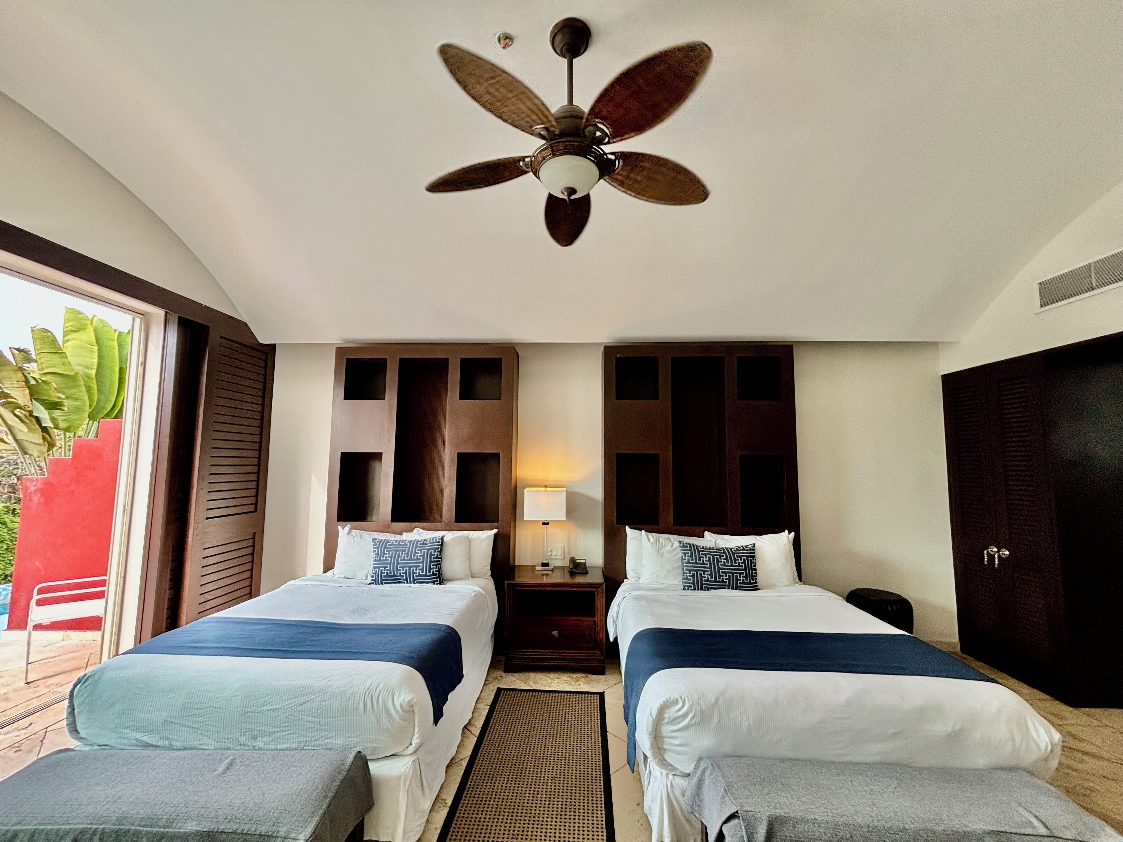 A hotel room with two beds, a ceiling fan, a nightstand with a lamp, and a view of the outdoors.