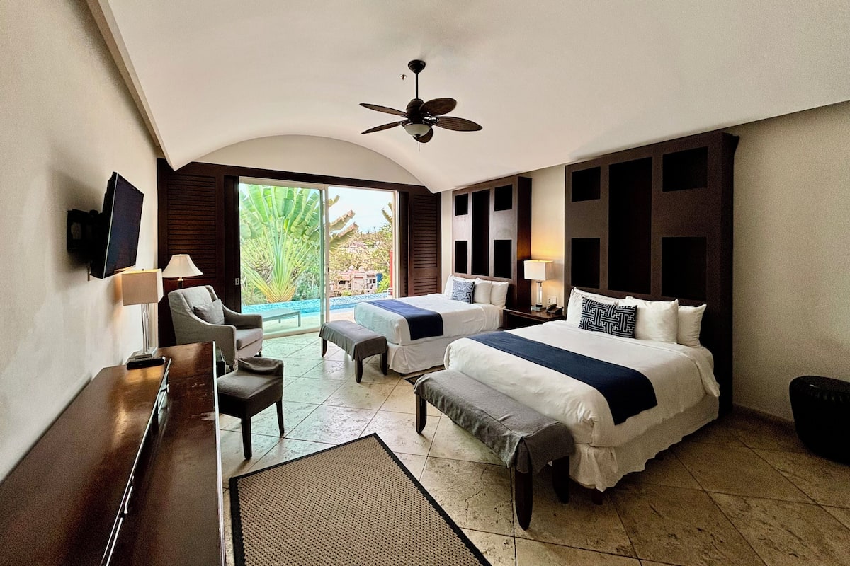 Une chambre d’hôtel avec deux lits, un bureau, une chaise, une petite table, un ventilateur de plafond et une grande fenêtre donnant sur un paysage verdoyant.