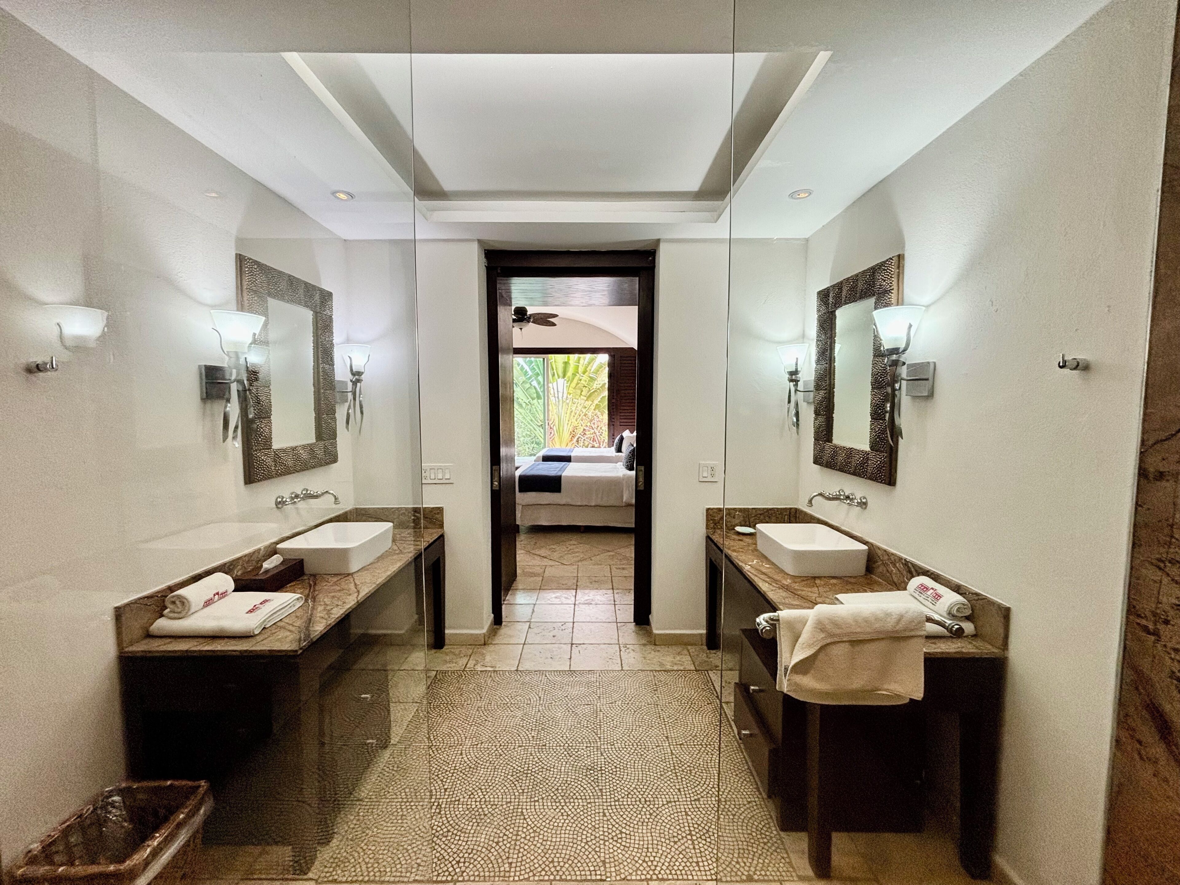 A modern bathroom with two sinks, a large mirror, and a view into a bedroom.