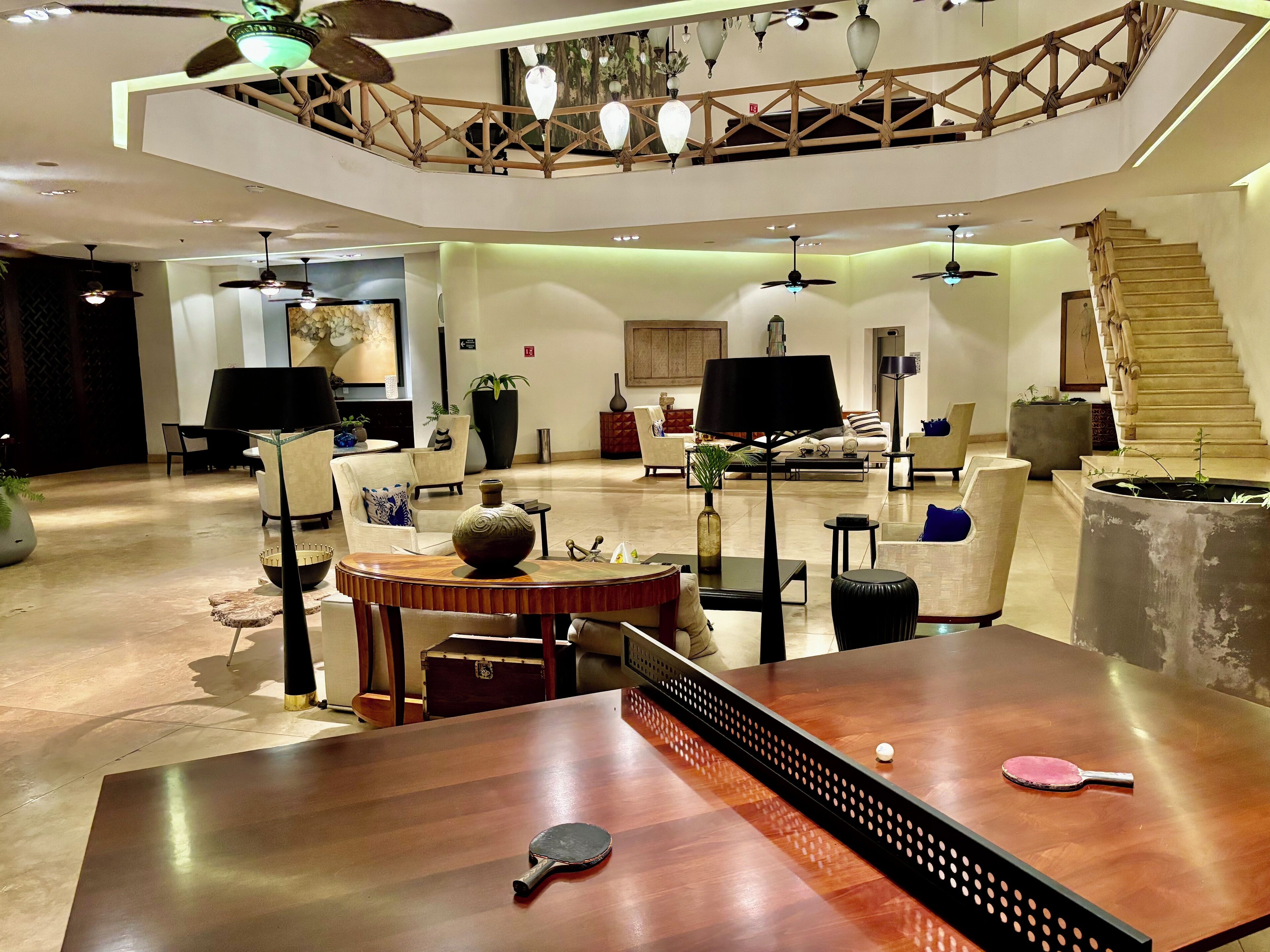 A spacious hotel lobby with a high ceiling, modern furniture, and a central wooden table.