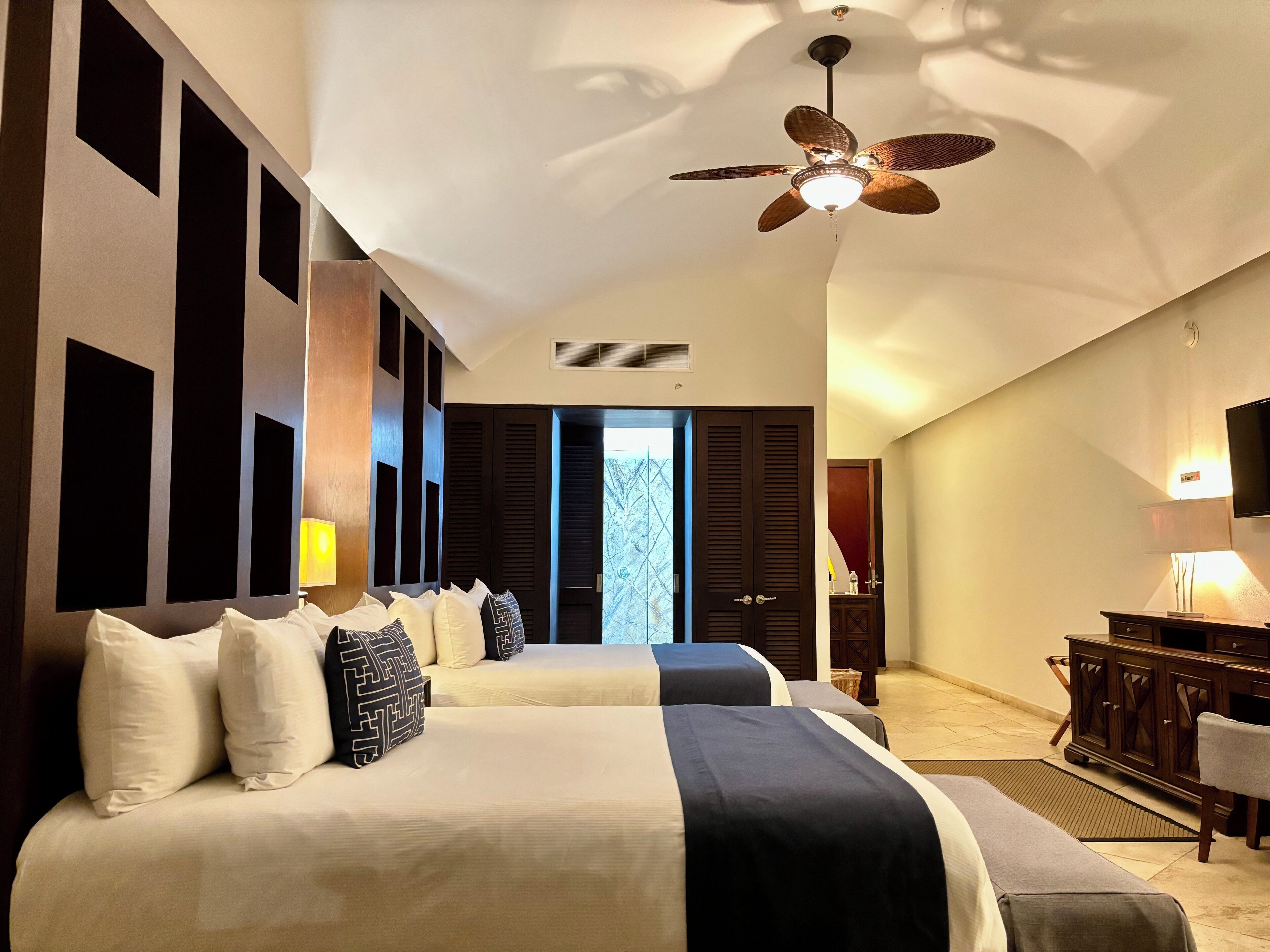 Habitación de hotel con dos camas, ventilador de techo, escritorio con lámpara y vista al exterior.