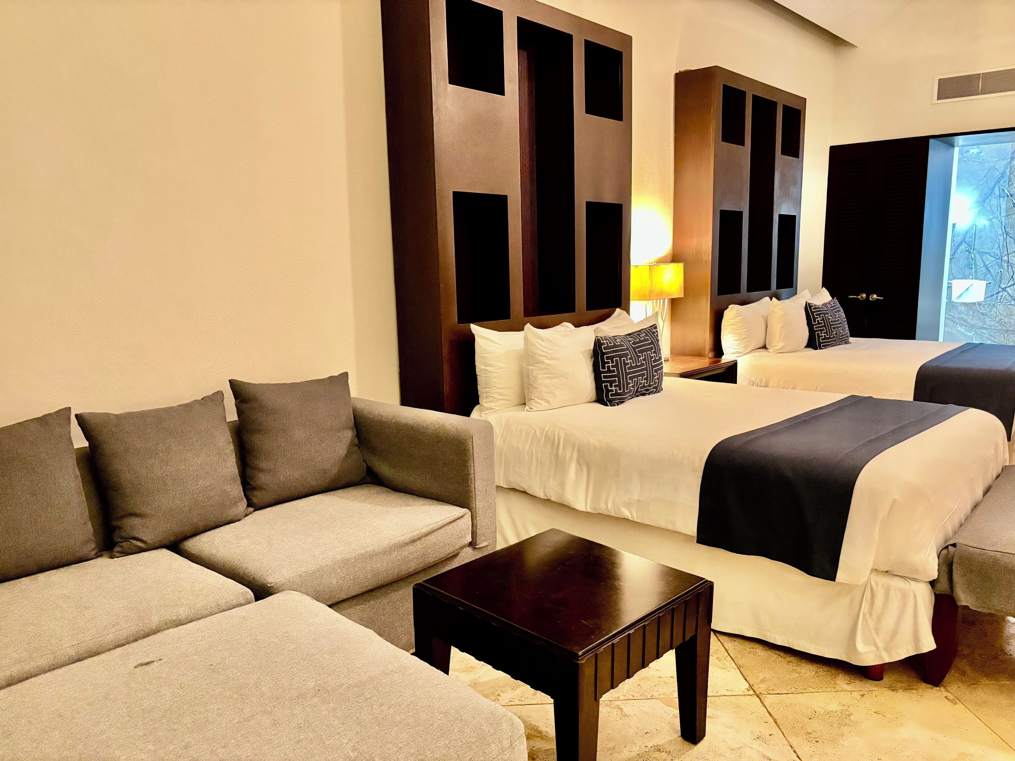 A hotel room with a sofa, two beds, a nightstand, and a lamp.