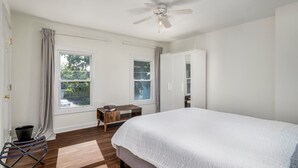 A bedroom with a large bed, two windows, a ceiling fan, and a small table.