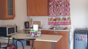 Una cocina compacta con armarios de madera, microondas, fregadero y refrigerador. Hay dos taburetes de bar y una pequeña isla con hervidor y botella de agua.