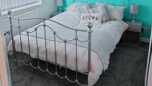Une chambre avec un lit en métal, un mur de couleur turquoise, une fenêtre avec des stores et un tableau encadré accroché au mur.
