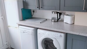 Une cuisine compacte avec des meubles gris, un lave-vaisselle blanc, un lave-linge à chargement frontal, une machine à café et un grille-pain.