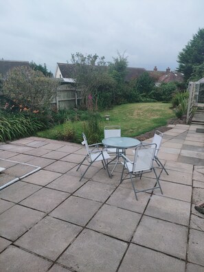A patio with a round table and four chairs, surrounded by a garden with various plants and a wooden fence. - Charming 2-Bed House in Fleetwood, England (Fleetwood)