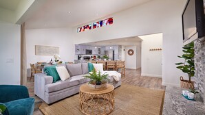 A spacious living room with a grey sofa, a round wicker coffee table, and a dining area with a kitchen in the background. - Oceanfront Gem: Dog-Friendly, AC, 2 Parking Spots, Walk to Pier & Shops (Oceanside)