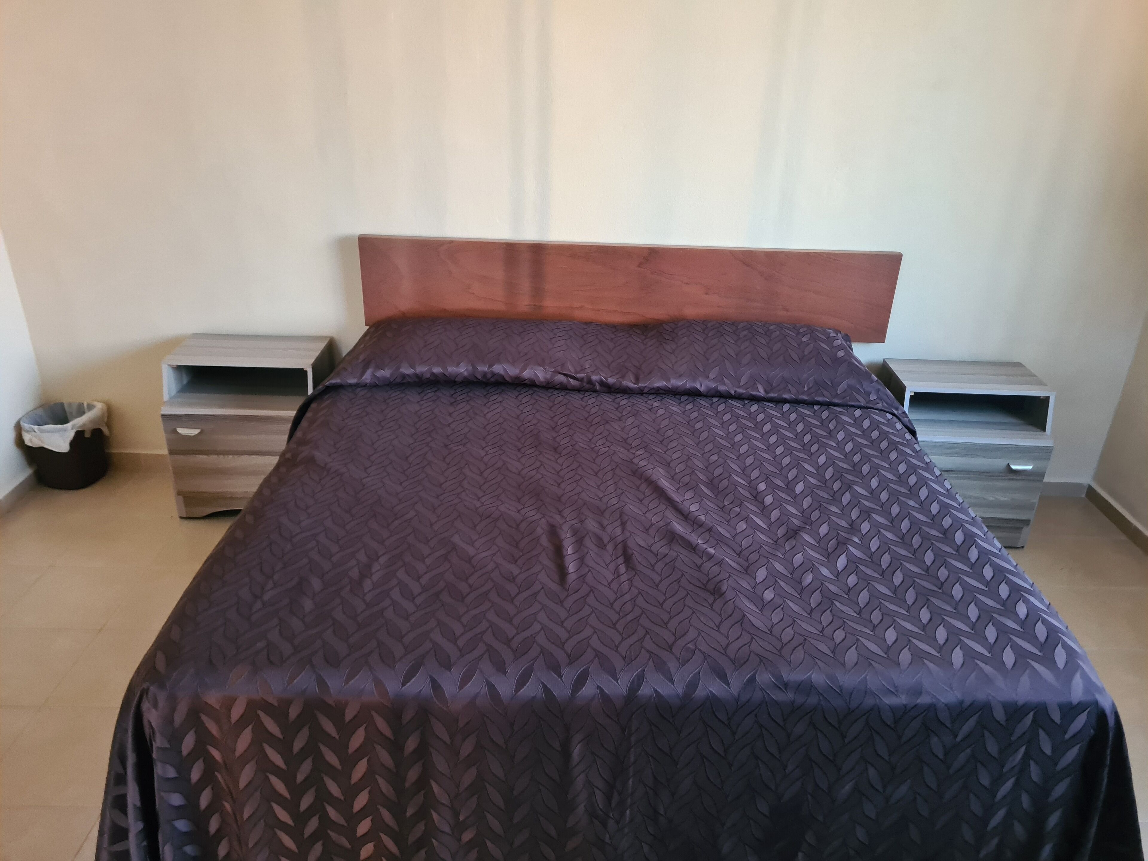 Una cama bien hecha con una colcha oscura estampada y un cabecero de madera. Hay dos mesitas de noche con cajones y un cubo de basura al lado.