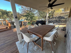 Eine überdachte Terrasse im Freien mit einem Holztisch und weißen Stühlen, einem Deckenventilator und Blick auf einen Hinterhof mit einem Grill.