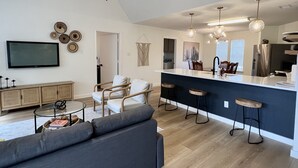 A modern living room with a grey sofa, wooden chairs, a glass coffee table, and a TV on the wall. - Scenic views on a quiet fishing cove. Boat Slip, Kayaks, Peddle Boat, SUPs. (Sunrise Beach Village)