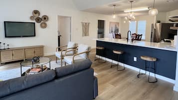 A modern living room with a grey sofa, wooden chairs, a glass coffee table, and a TV on the wall.
