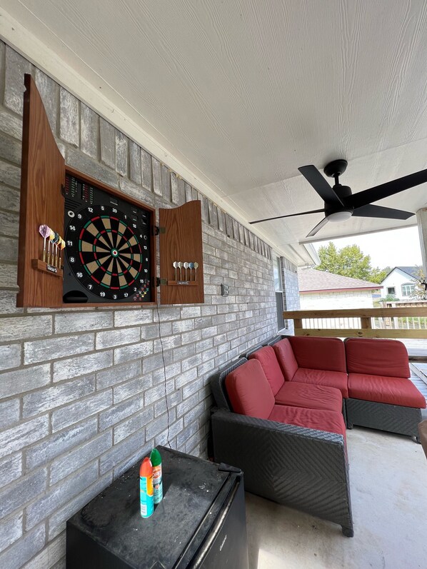 Een overdekt terras met een dartbord bevestigd aan een bakstenen muur, een plafondventilator en een hoekbank met rode kussens.