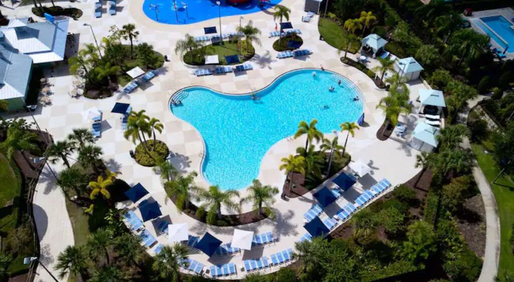 En vy från ovan av ett semesterområde med en stor swimmingpool omgiven av palmer och solstolar.