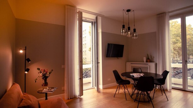 Ein modernes Wohnzimmer mit Sofa, Esstisch und Stühlen, einem flachbildfernseher an der Wand und einem Balkon mit Stadtblick.
