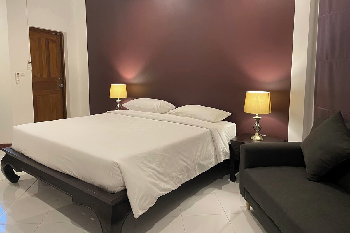 A bedroom with a bed, two bedside lamps, a dark-colored sofa, and a door.