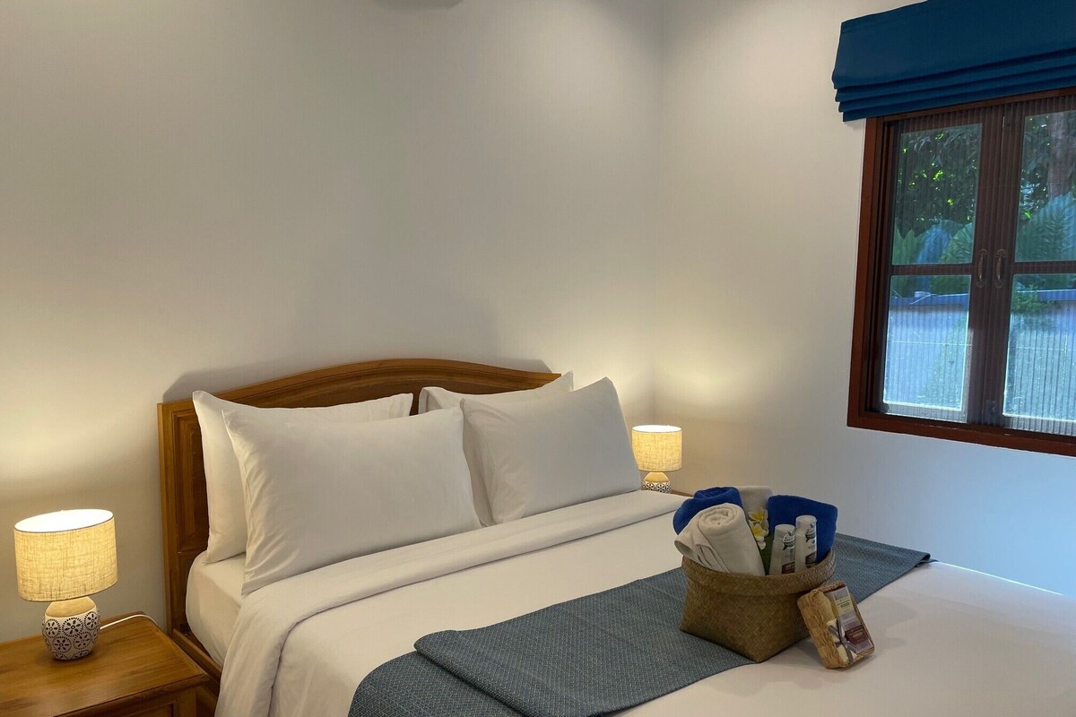 A neatly made bed with white linens, two bedside lamps, a window with a view of greenery, and a small basket with toiletries.