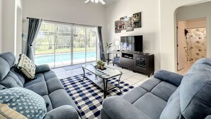 Un salon moderne avec un canapé gris, une table basse et un téléviseur à écran plat. On y trouve une grande fenêtre avec des rideaux, un ventilateur de plafond et une porte ornée d’un motif floral.