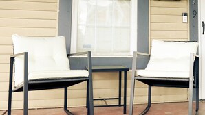 A porch with two white chairs and a small table, a window with blinds, and a mailbox.