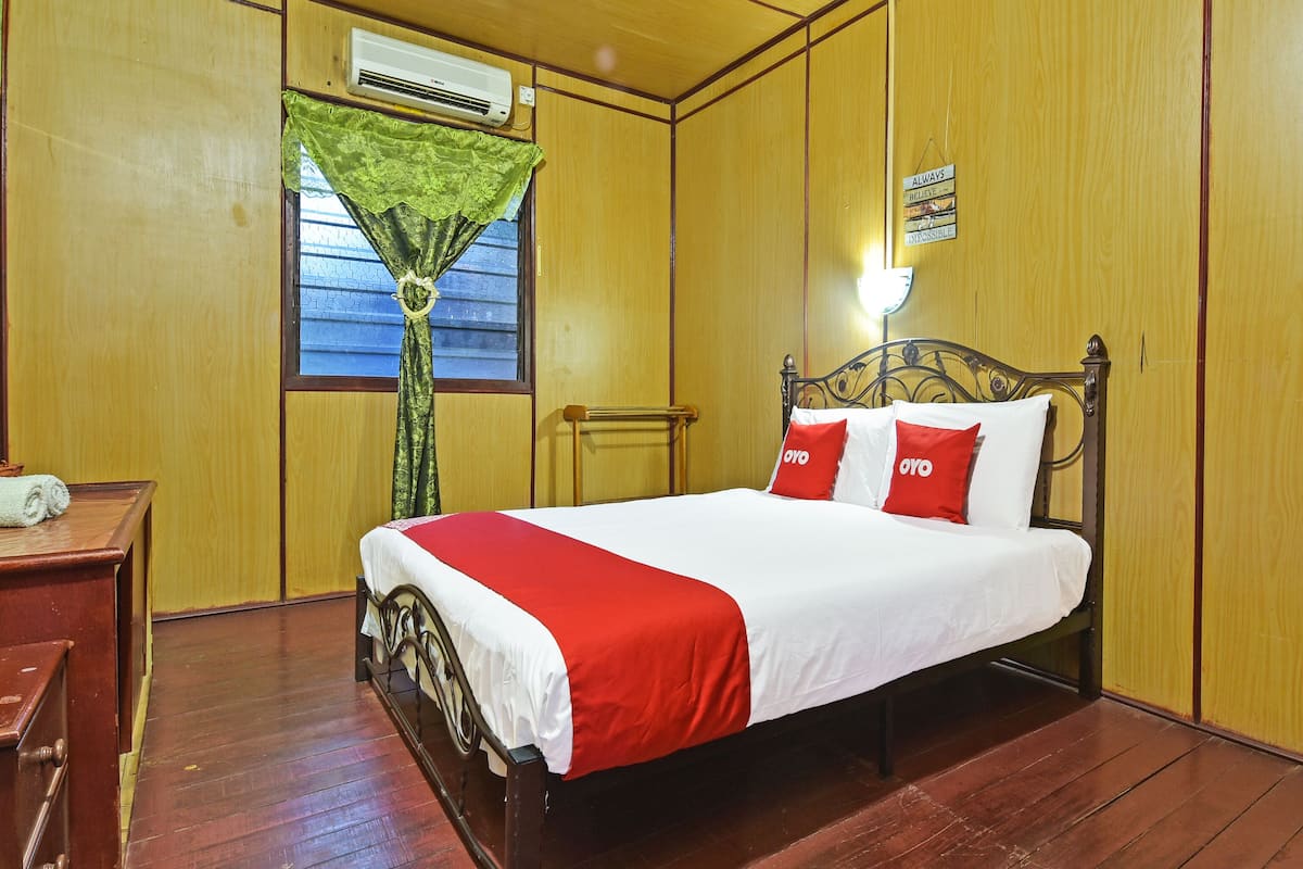 Un lit bien fait, recouvert d’une couvre-lit rouge et blanche et agrémenté de deux oreillers rouges, une table de chevet en bois avec un téléphone, un plancher en bois et une fenêtre avec un rideau vert.