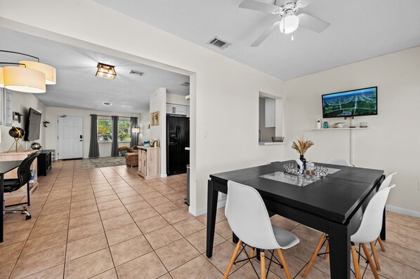 A modern kitchen with a black refrigerator, a dining area with a black table and white chairs, and a TV mounted on the wall.