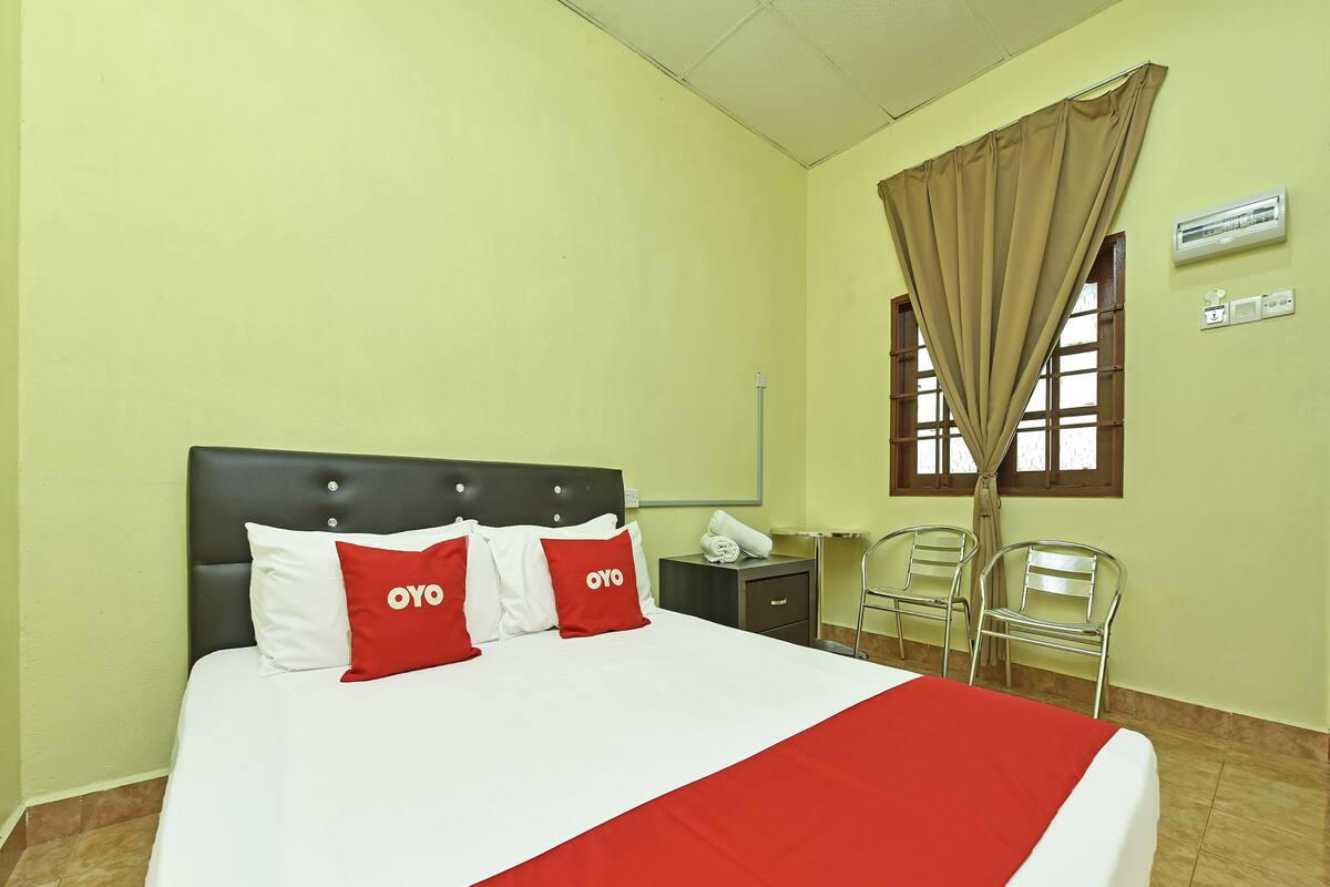 A hotel room with a bed, two red pillows with "OYO" branding, a bedside table, a chair, a window with curtains, and a wall-mounted air conditioner.