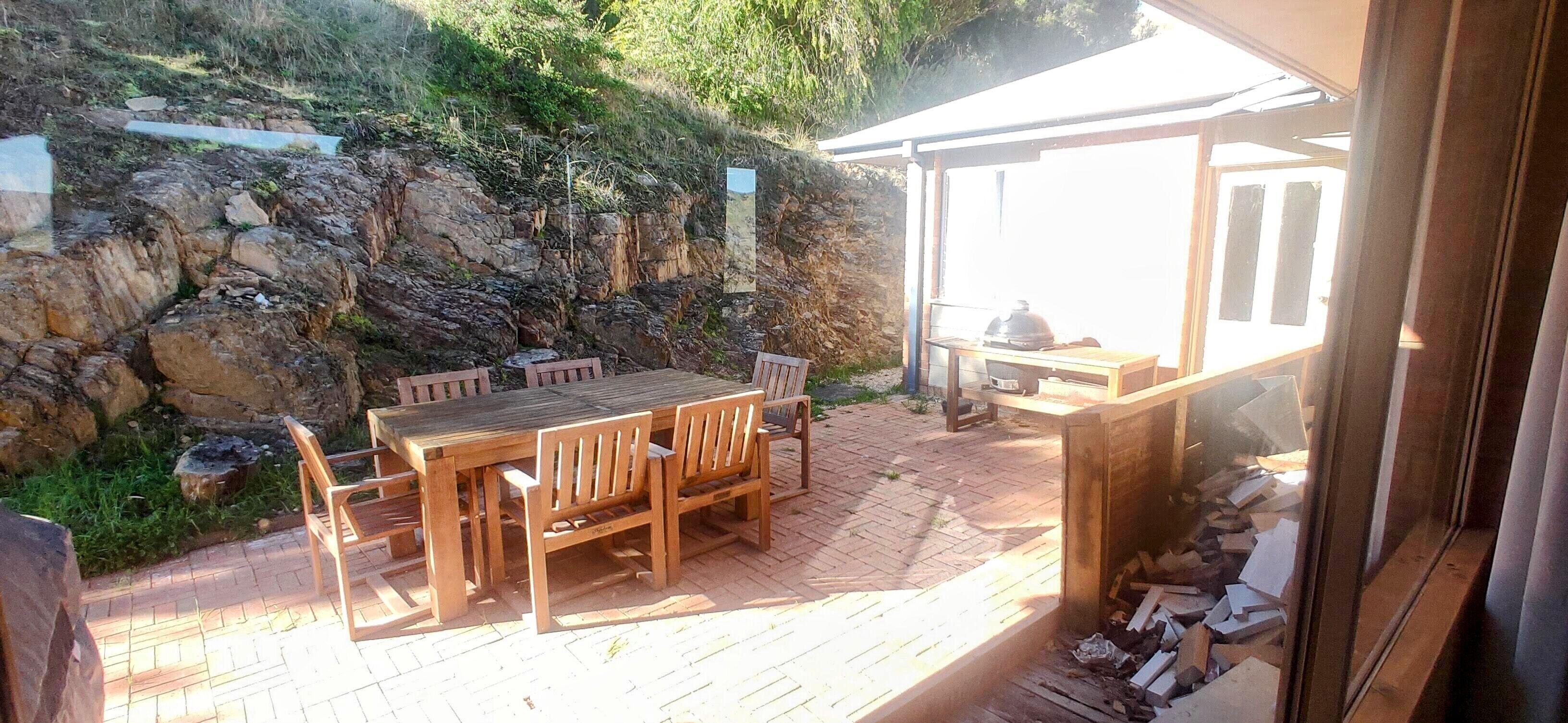 An outdoor patio with a wooden table and chairs, a barbecue grill, and a stone wall.