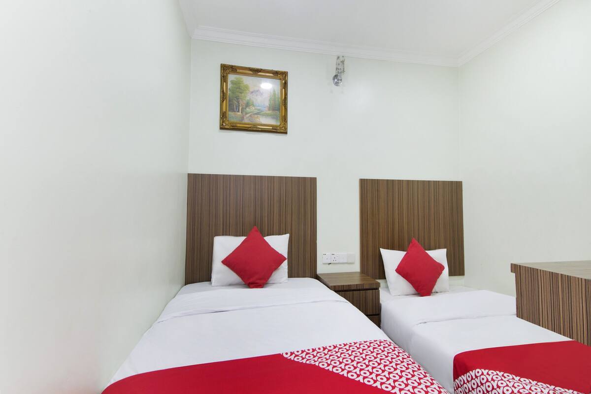 A hotel room with two beds, each with a wooden headboard, white bedding, and red pillows. A painting hangs on the wall above the beds.