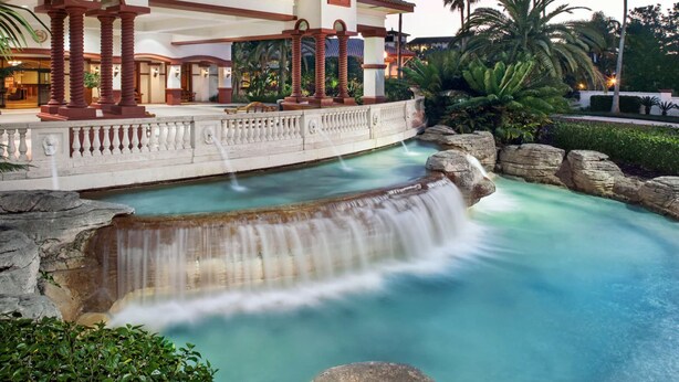 Un hall d’hôtel agrémenté d’un élément décoratif aquatique, de colonnes et offrant une vue sur la verdure environnante.