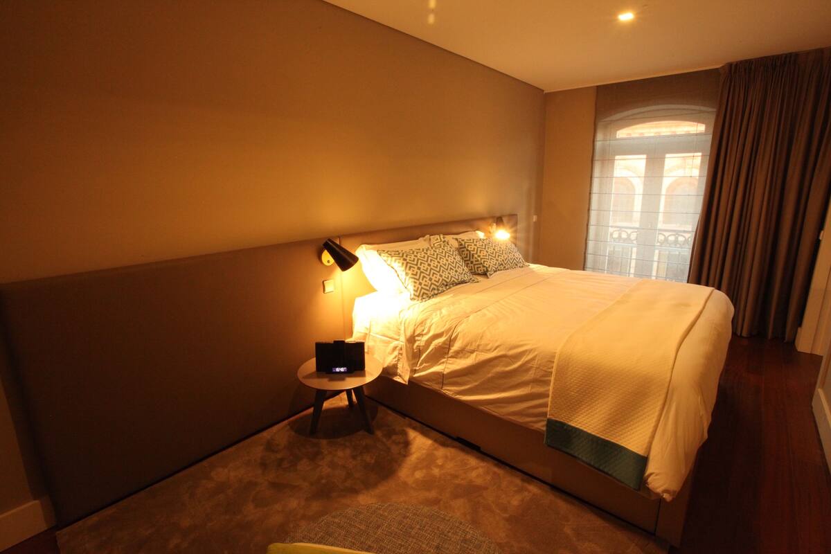 Ein Hotelzimmer mit einem großen Bett, einem Nachttisch mit Lampe und Blick auf ein Gebäude durch ein Fenster.