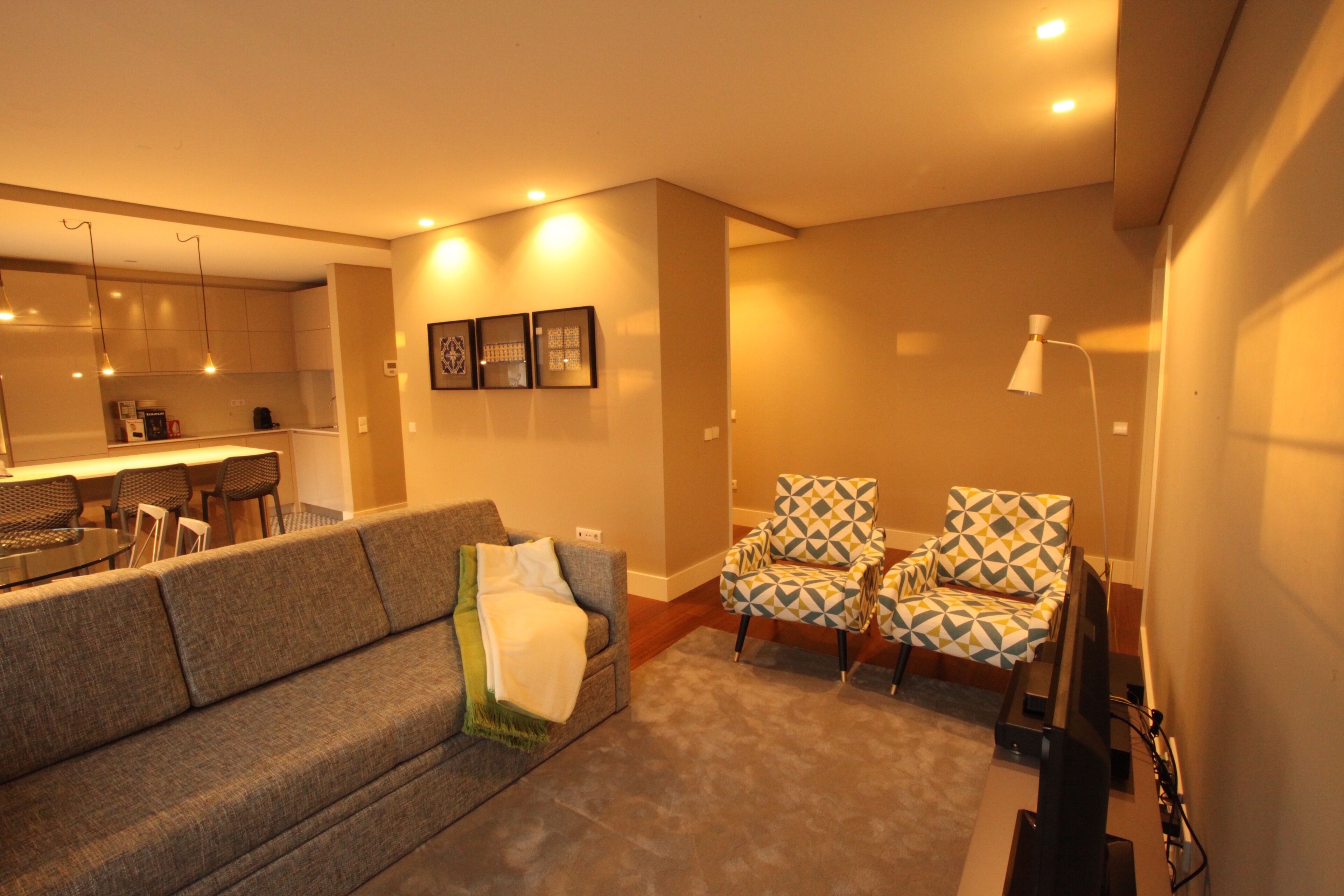 A living room with a sofa, two patterned armchairs, a coffee table, and a TV. There is a kitchen in the background with a dining area.