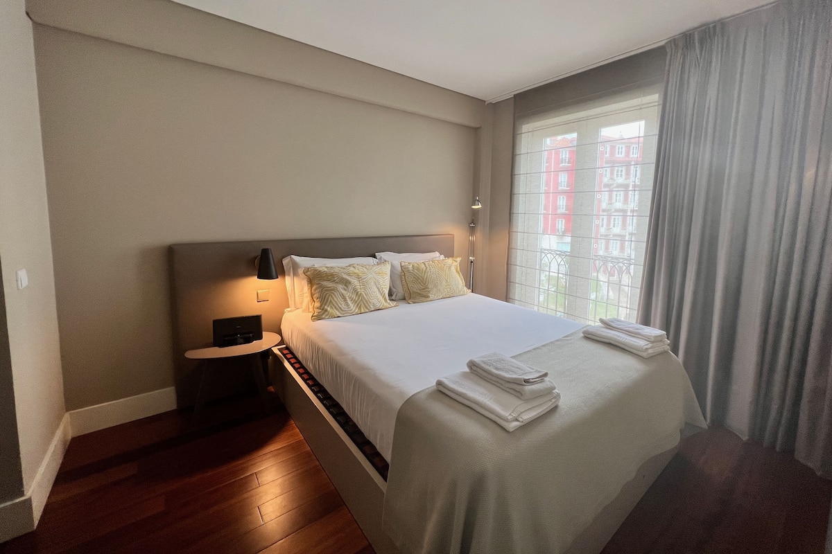 Ein Hotelzimmer mit einem großen Bett, einem Nachttisch, einer Lampe und Blick auf Gebäude durch das Fenster.